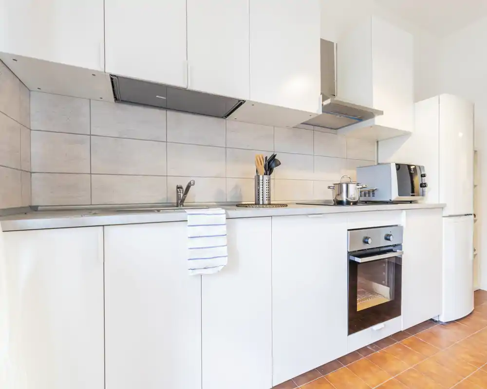Cucina moderna e luminosa con mobili bianchi, forno incassato, frigorifero e piano di lavoro pulito; stile ordinato con portautensili e asciughino.