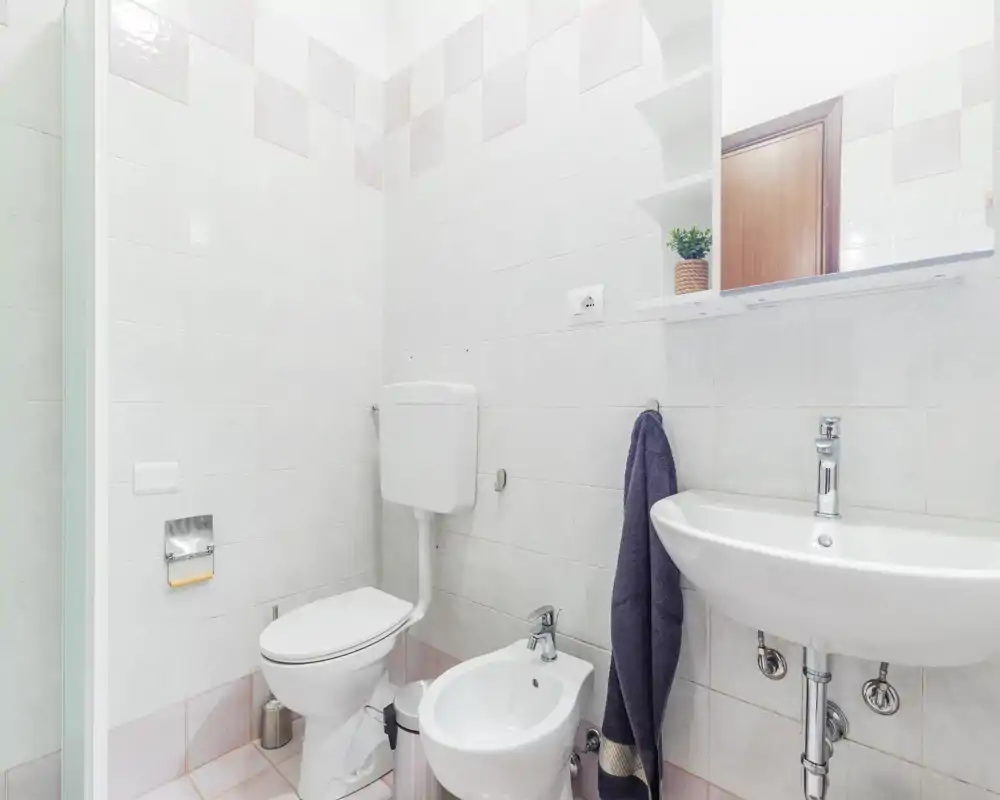 Bagno luminoso e pulito con WC, bidet e lavabo a parete, piastrelle alle pareti e un pensile con specchio sopra il lavabo.
