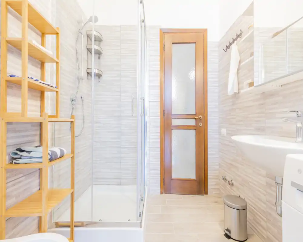 Bagno moderno e luminoso con box doccia in vetro, lavabo a parete, scaffalatura in legno e piastrelle neutre, ambiente pulito e accogliente.
