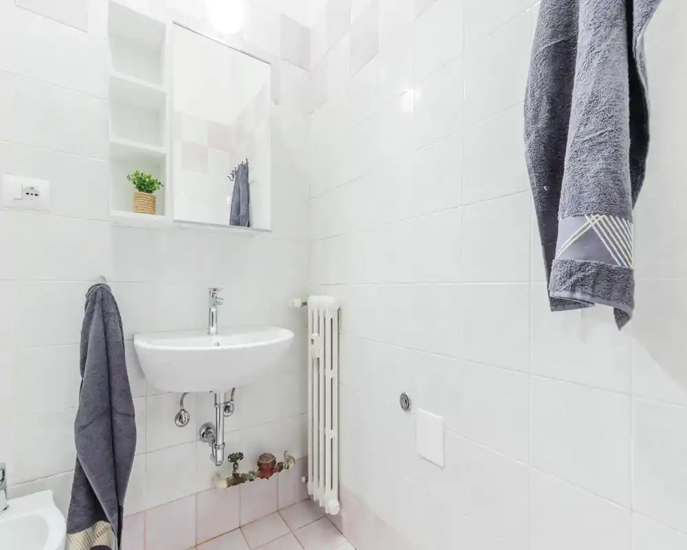 Bagno compatto e luminoso con piastrelle bianche, lavabo a parete, termosifone e asciugamani appesi; ambiente pulito e ben illuminato, adatto agli annunci.