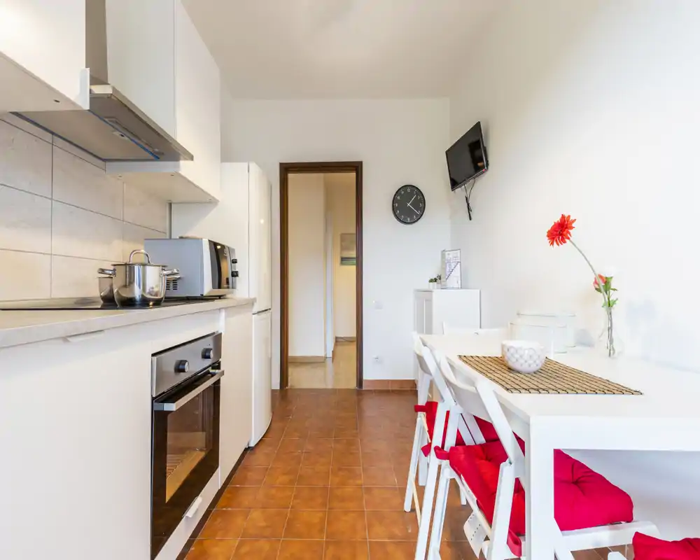 Cucina moderna e luminosa con forno integrato, frigorifero e un piccolo angolo pranzo con sedie con cuscini rossi e vaso decorativo semplice.