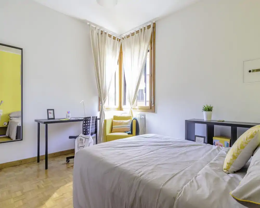 Bright, modern bedroom with a comfortable double bed, soft neutral linens, a small desk and a cosy yellow armchair by the window letting in abundant natural light.