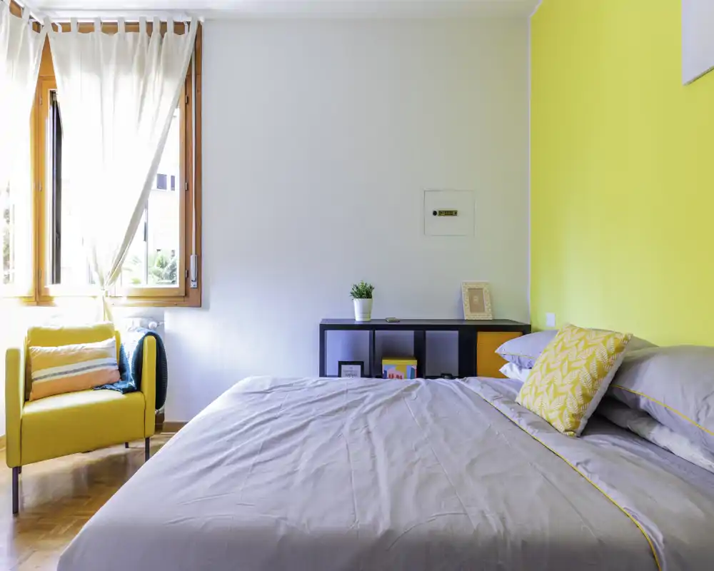 Bright, modern bedroom with a large bed dressed in neutral linens, a yellow accent wall and a cozy yellow armchair by the window letting in abundant natural light.