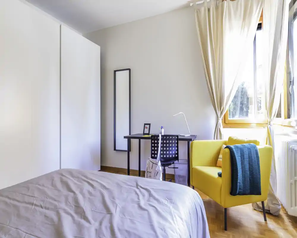 Bright, modern bedroom with a large bed in the foreground, a work desk and a cheerful yellow armchair by the window that fills the room with natural light.