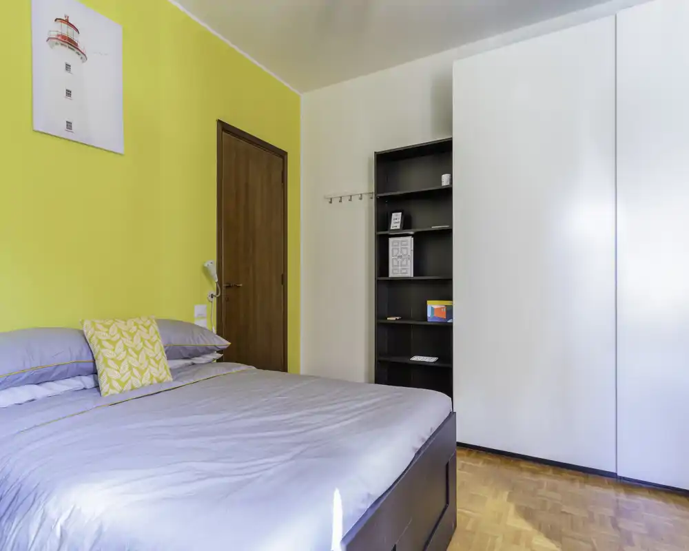 Bright, neatly staged bedroom with a large bed, a yellow accent wall and built-in wardrobe; natural light and simple decor create a fresh, welcoming space.