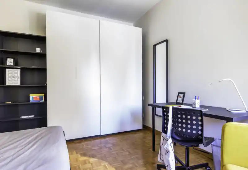 Modern, bright bedroom with a large white wardrobe, a study desk and a partially visible bed. Clean, minimalist styling with parquet floors and a comfortable workspace near a full-length mirror.