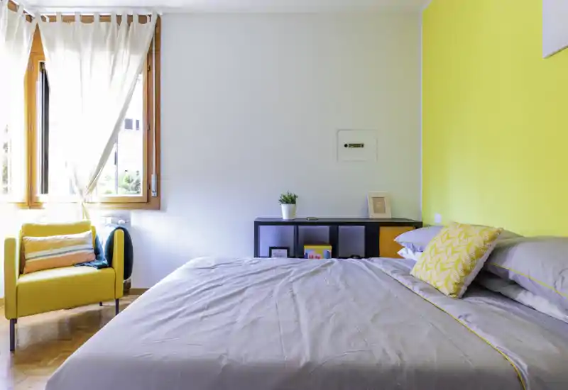 Bright, modern bedroom with a large bed dressed in neutral linens, a yellow accent wall and a cozy yellow armchair by the window letting in abundant natural light.