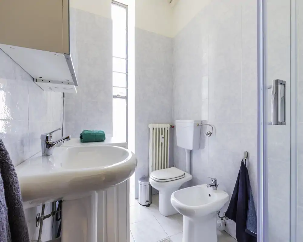 Clean, bright bathroom with a pedestal sink, toilet, bidet and a glass shower door; neutral tiled walls and a tall window provide natural light.
