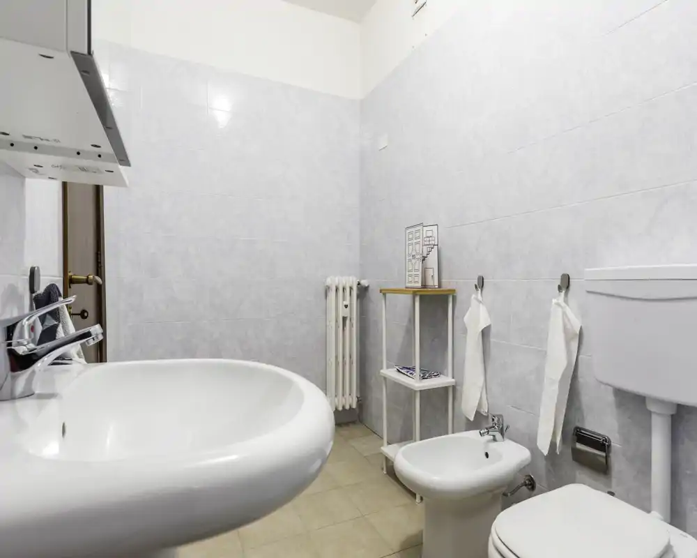 Clean, well-lit bathroom with a sink, bidet, toilet and simple shelving; neutral tiles and uncluttered layout create a fresh, functional space.