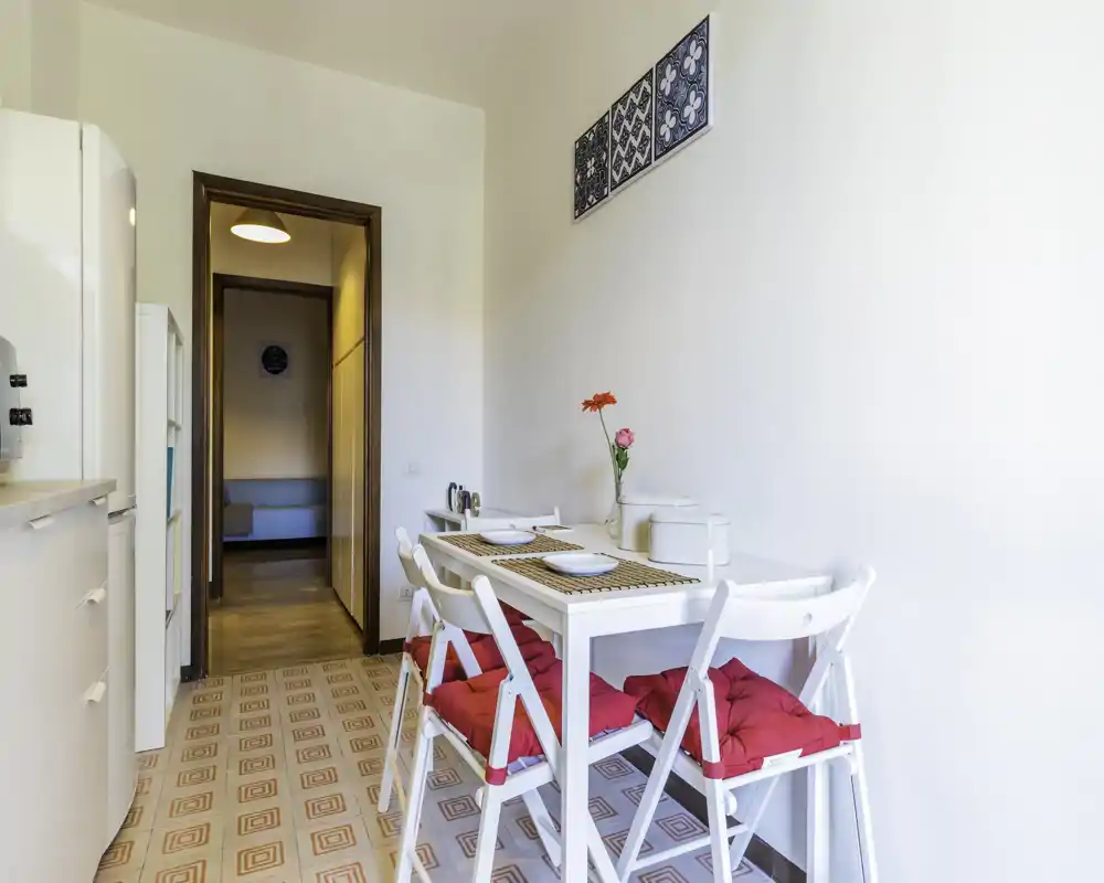 Compact, bright dining corner with a white table and four chairs topped with red cushions, placemats and a simple floral centerpiece — ideal for cozy meals.