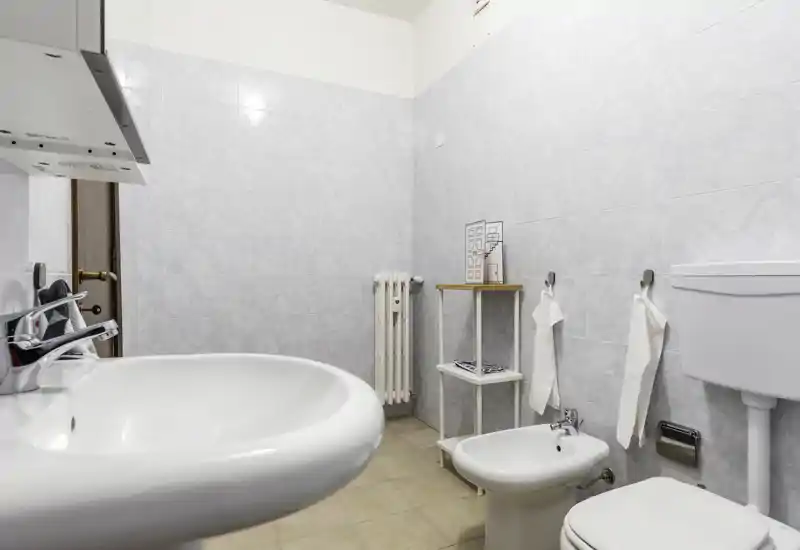 Clean, well-lit bathroom with a sink, bidet, toilet and simple shelving; neutral tiles and uncluttered layout create a fresh, functional space.