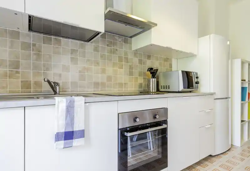 Modern compact kitchen with white cabinets, built-in oven, stovetop and refrigerator. Bright, neatly styled workspace with tiled backsplash and small appliances.