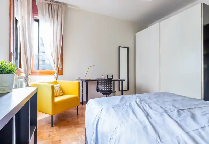 Camera da letto moderna e luminosa con comodo letto matrimoniale, poltrona gialla vicino alla finestra, scrivania e grande armadio — fresca, ben illuminata e accogliente.