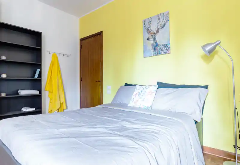 Camera da letto luminosa e accogliente con letto matrimoniale ben rifatto su una parete gialla, cuscino decorativo e lampada da terra. Scaffale e appendiabiti offrono spazio per riporre, perfetta per affitti brevi.