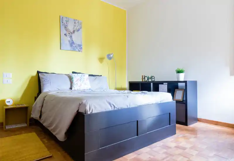 Camera da letto moderna e luminosa con un grande letto dalla struttura scura davanti a una parete gialla. L'ambiente presenta biancheria morbida, cuscino decorativo, contenitori vicino al letto e una lampada da terra, creando una soluzione accogliente e ben illuminata ideale per annunci.