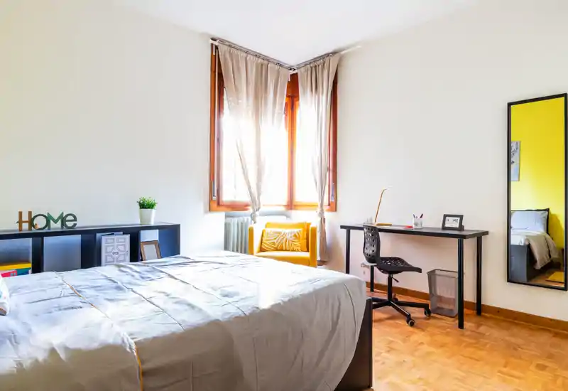 Camera luminosa e moderna con un ampio letto in primo piano, una comoda poltrona gialla vicino alla finestra e una scrivania funzionale — perfetta per soggiorni confortevoli e smart working.