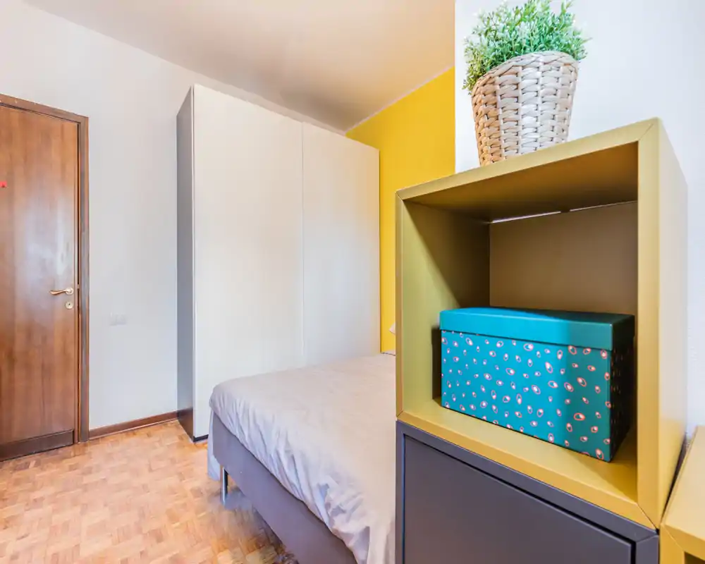 Compact bedroom with a neatly made double bed, a large white wardrobe and a styled bedside shelf featuring a decorative box and plant. The room shows warm parquet flooring and a yellow accent wall, offering a bright, inviting atmosphere.