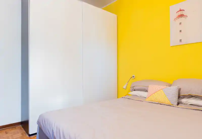 Bright, modern bedroom with a large bed dressed in neutral linens, a decorative striped cushion and a striking yellow accent wall — clean lines and simple furnishings create a fresh, welcoming space.