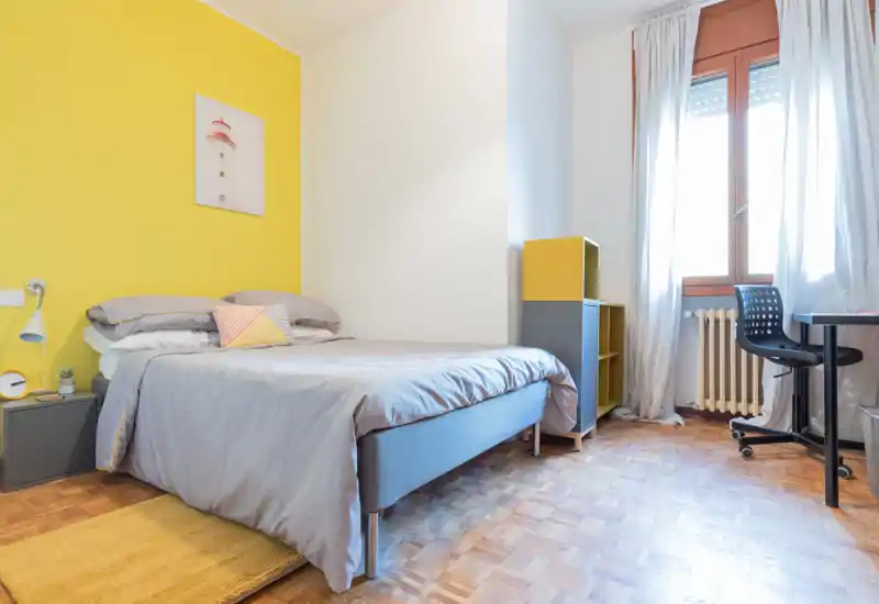 Bright, neatly staged bedroom with a full-size bed against a yellow accent wall, soft grey bedding, a bedside lamp, small storage units and a desk by the window — ideal for a single occupant or couple.
