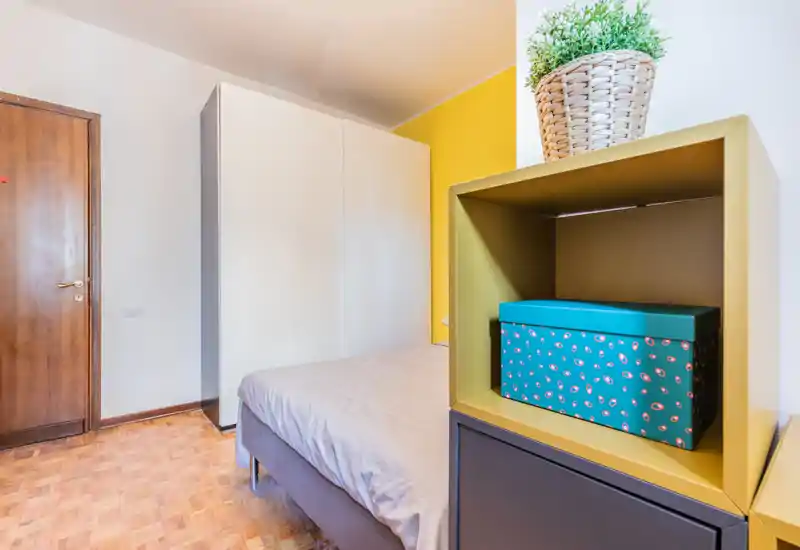 Compact bedroom with a neatly made double bed, a large white wardrobe and a styled bedside shelf featuring a decorative box and plant. The room shows warm parquet flooring and a yellow accent wall, offering a bright, inviting atmosphere.