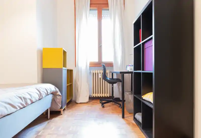 Bright, tidy single bedroom with a visible bed to the left, compact desk by the window and modular shelving for storage — ideal for a student or single occupant.