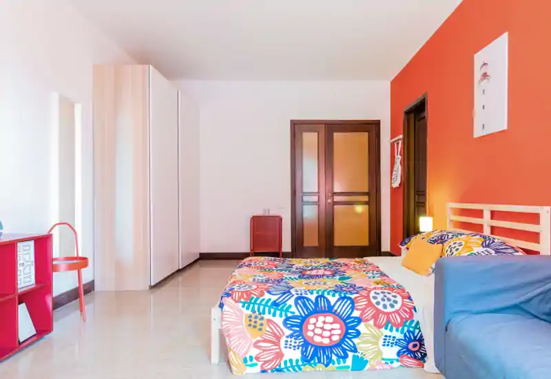 Bright, colorful bedroom with a patterned floral duvet, wooden headboard and a small sofa at the foot of the bed. The room features a large wardrobe, warm orange accent wall and neat, well-lit styling ideal for rental photos.