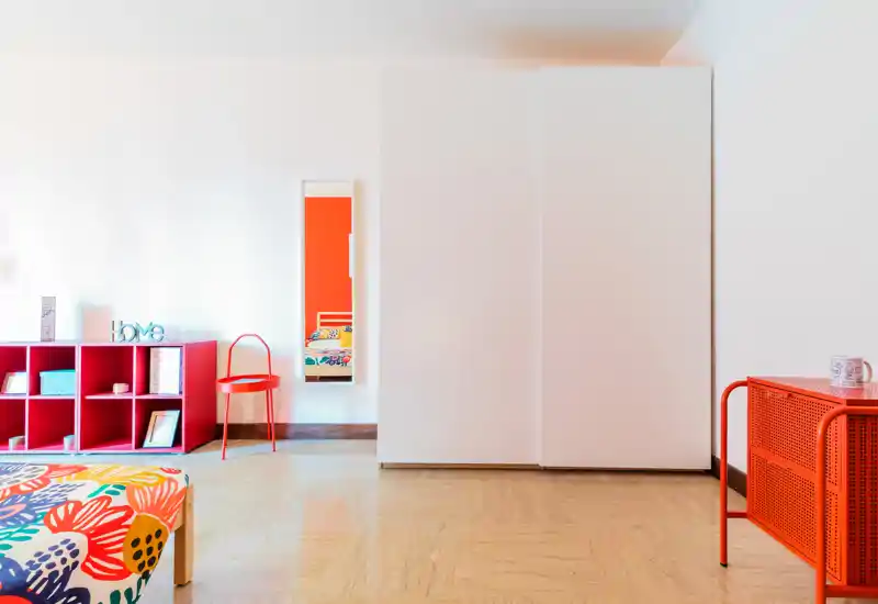 Bright, modern bedroom corner with a colorful patterned bedspread, white wardrobe and red-accent storage and chair; contemporary, cheerful styling ideal for a vibrant rental listing.