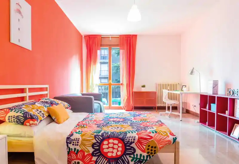 Bright, colorful bedroom with a full bed in the foreground, patterned bedding, a small sofa, desk and balcony window offering natural light — cheerful and well-styled for rental photos.