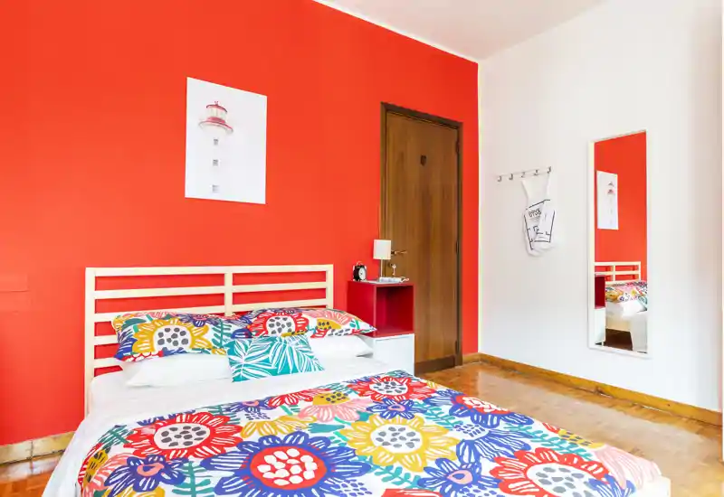 Bright, colorful bedroom with a wooden slatted bed, patterned bedding and a bold red accent wall — well-lit and cheerfully styled for a cozy stay.
