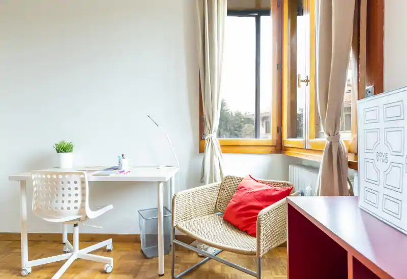 Bright, cozy living nook with a rattan armchair, red accent cushion and a compact desk by a large window—ideal for working or relaxing with plenty of natural light.