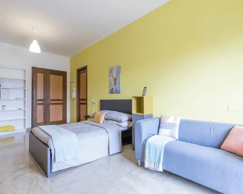 Camera da letto luminosa e moderna con letto matrimoniale, biancheria morbida e un comodo divano accanto — parete accentuata gialla e contenitori ordinati per un ambiente arioso e accogliente.