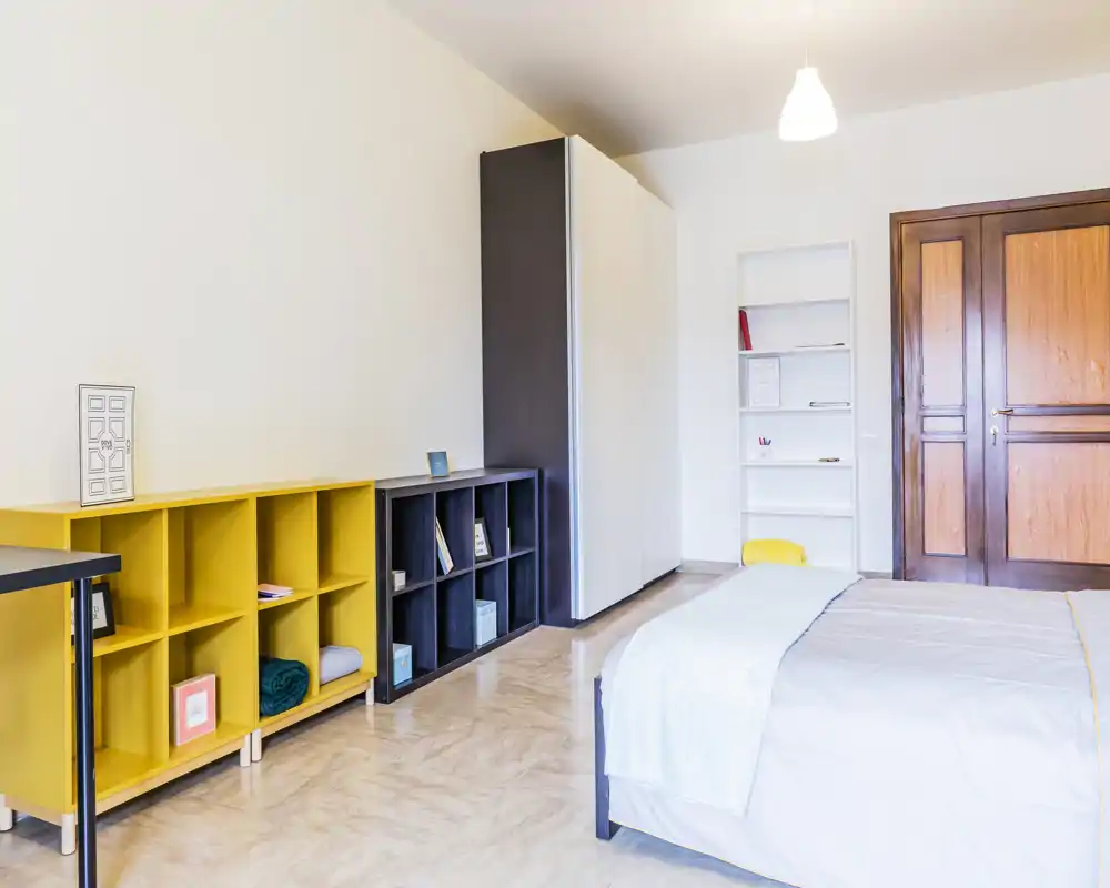 Camera da letto luminosa e minimal con letto matrimoniale ben rifatto, scaffalature moderne e un ampio armadio. L'ambiente è arioso e accogliente grazie alle pareti chiare e alla buona illuminazione.