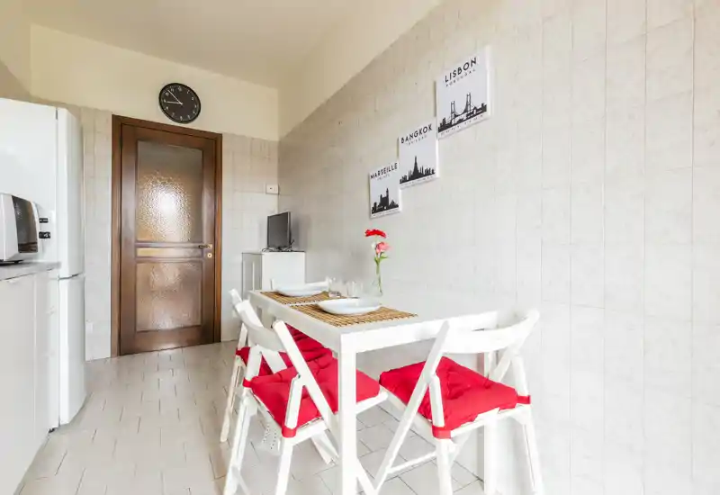 Bright, tidy kitchen with a small dining table set for two. White furniture and red seat cushions create a fresh, modern look ideal for short-term rentals.