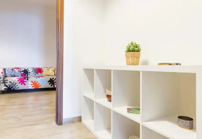 Bright, minimal living area seen from an entry corner: a white cubed shelf in the foreground and a colorful sofa visible through the doorway suggest a modern lounge space.