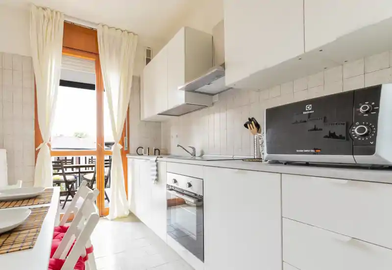 Bright, modern kitchen with white cabinetry, built-in oven and microwave, and a small dining area opening onto a sunny balcony.