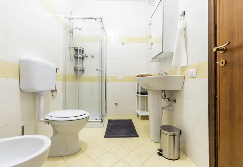 Bright, well-lit bathroom with shower cabin, pedestal sink, toilet and bidet on tiled floor; modern fixtures and neutral tones create a clean, functional space.