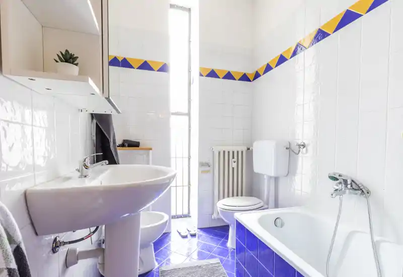 Bright, well-maintained bathroom with a pedestal sink, toilet, bathtub and blue tiled floor. Tall window provides abundant natural light for a fresh atmosphere.