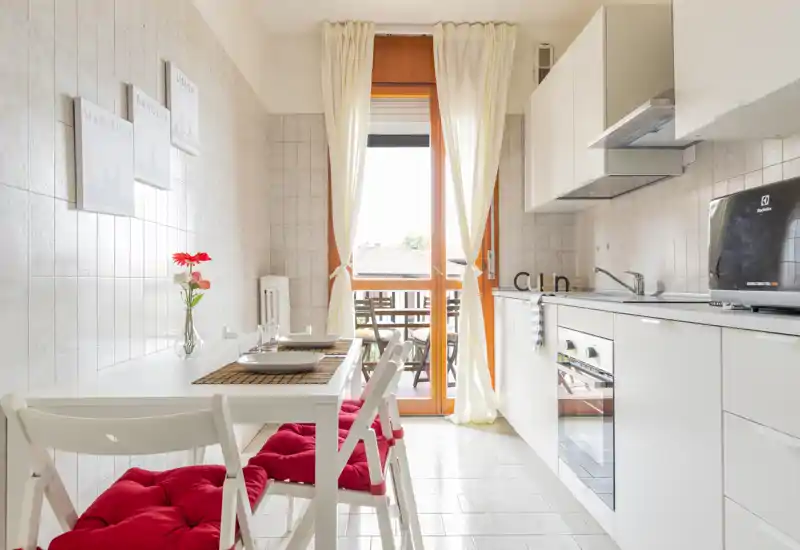 Bright, modern kitchenette with white cabinets, built-in oven, and a dining table set for two opening onto a small balcony — ideal for cozy city stays.