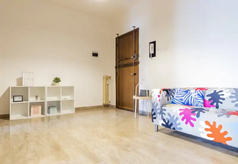 Bright, minimal living room with a colorful patterned sofa, light marble floor and simple shelving — clean and modern space ready for guests.