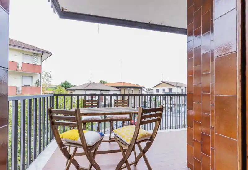 Sunny balcony with a small wooden table and two chairs, colorful cushions and railings overlooking neighboring houses — ideal for morning coffee.