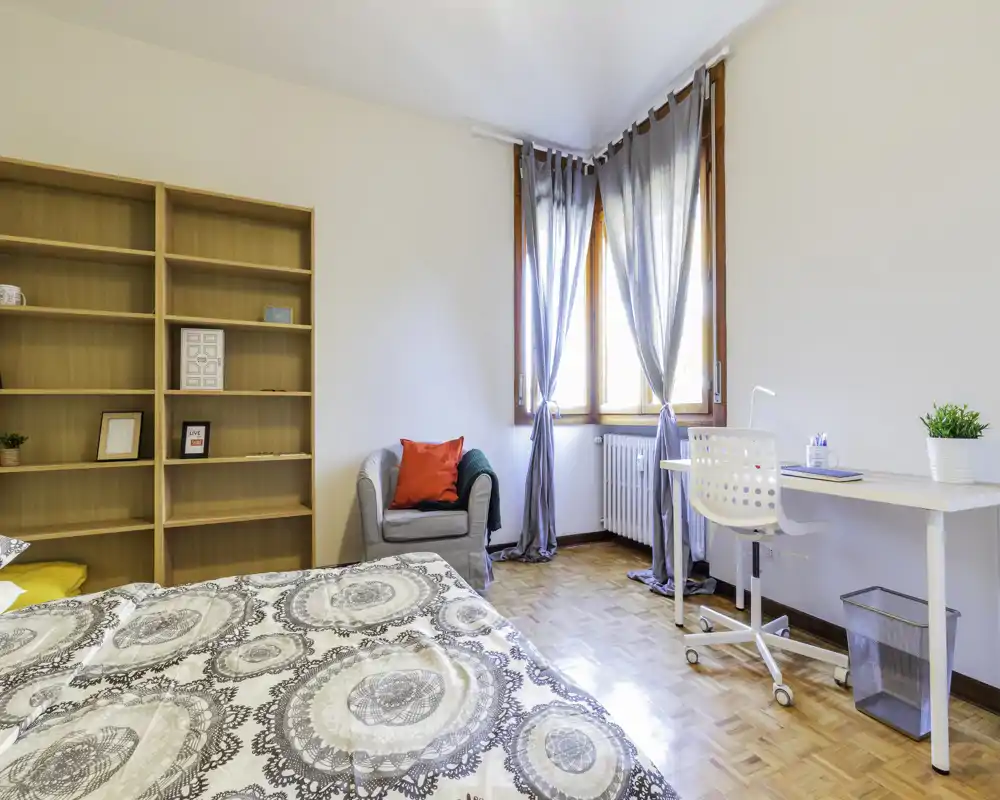 Dormitorio luminoso y bien decorado con la cama en primer plano, un sillón acogedor, estantería y escritorio junto a la ventana que aporta mucha luz natural.