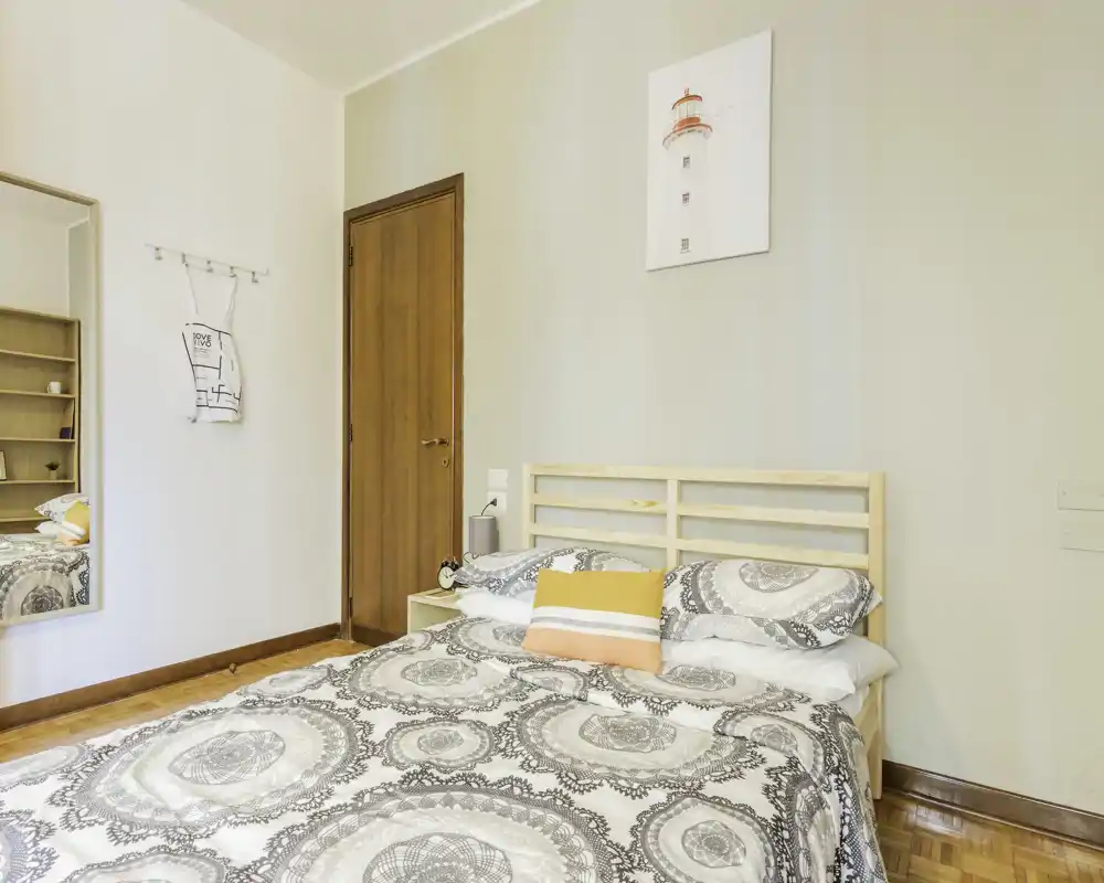 Dormitorio luminoso y ordenado con cabecero de madera, edredón estampado y cojines decorativos; suelo de parqué y una decoración de pared sencilla crean ambiente acogedor.