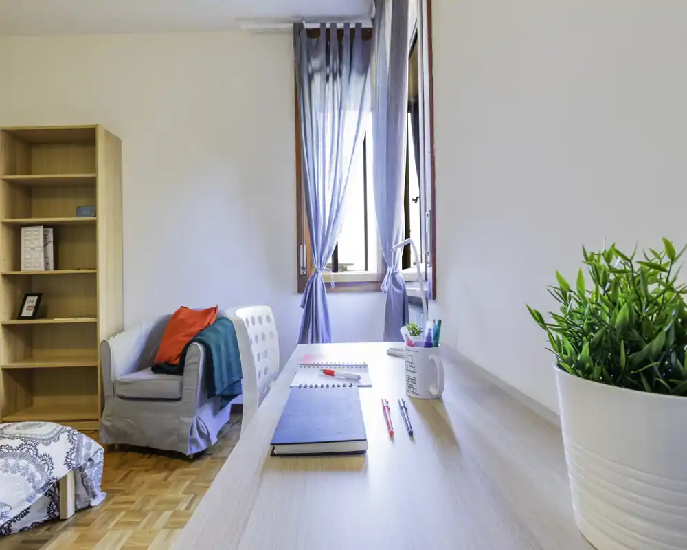 Dormitorio luminoso y ordenado con sillón, ventana abierta con cortinas y un escritorio en primer plano — ideal para una persona o estudiante.