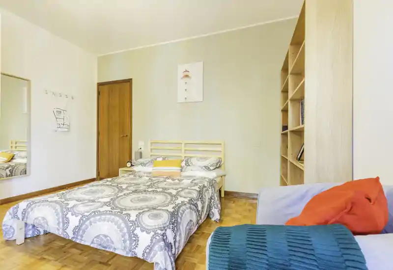 Dormitorio luminoso y ordenado con cama doble, edredón estampado y mesita de noche. Una estantería y un pequeño sofá con cojines de colores completan este espacio acogedor y bien iluminado, ideal para el anuncio.