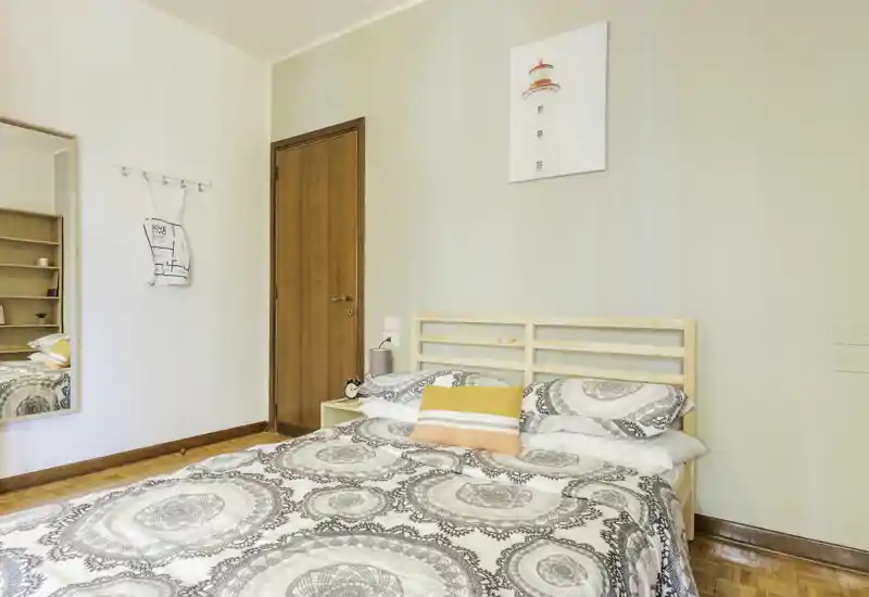 Dormitorio luminoso y ordenado con cabecero de madera, edredón estampado y cojines decorativos; suelo de parqué y una decoración de pared sencilla crean ambiente acogedor.
