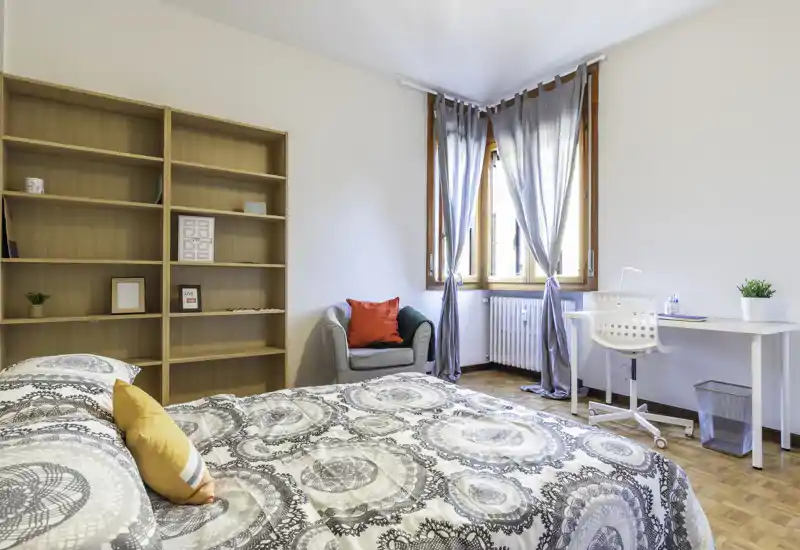 Dormitorio luminoso y bien decorado con una cama doble estampada en primer plano, un sillón cómodo con cojines de color, una gran estantería y una zona de trabajo junto a la ventana.