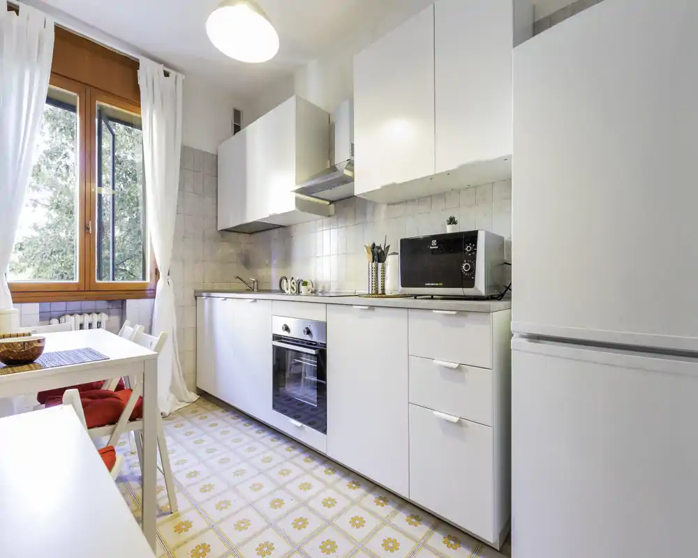 Cocina moderna y luminosa con armarios blancos, horno empotrado, microondas y mesa junto a una ventana grande. El suelo con azulejos de motivos cálidos y la luz natural crean un espacio acogedor para cocinar y comer de forma informal.