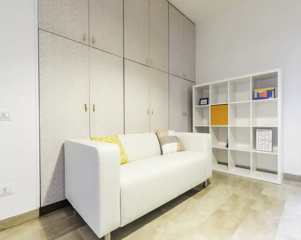 Pequeña sala de estar moderna y luminosa con sofá blanco, cojines decorativos y estantería cuadriculada; suelo efecto mármol y armarios empotrados ofrecen un aspecto ordenado y contemporáneo.