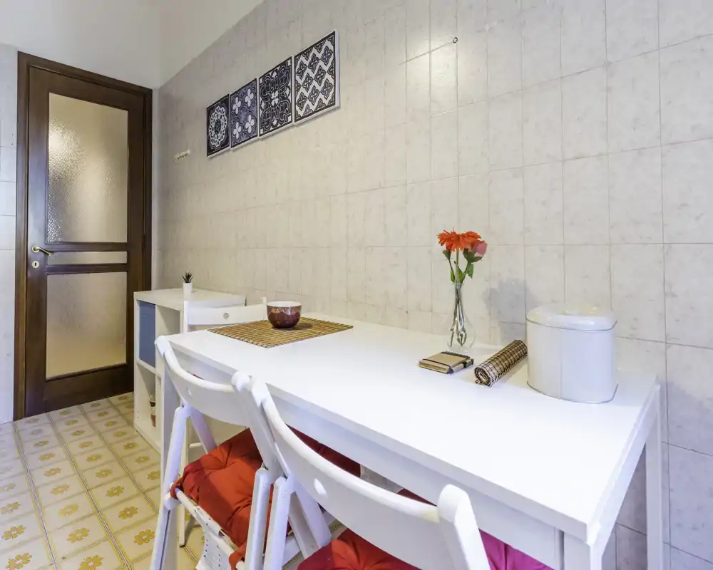Acogedor rincón comedor luminoso con mesa blanca, dos sillas con cojines rojos, azulejos decorativos y un jarrón con flores — ideal para comidas informales.