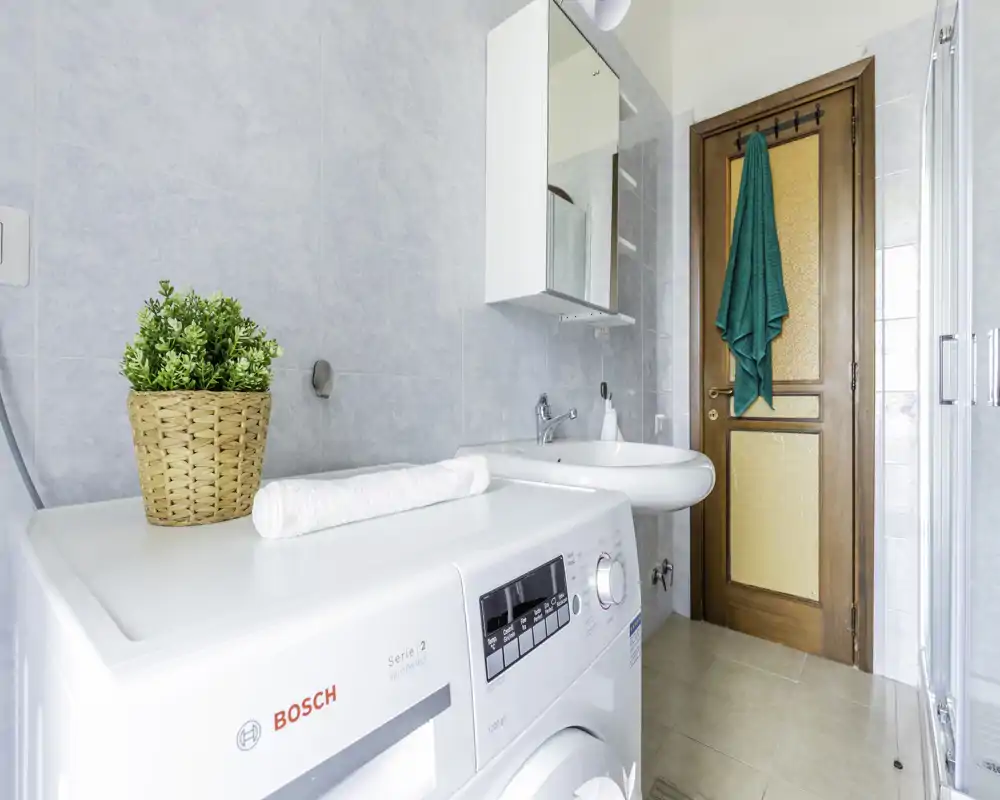 Baño compacto y luminoso con lavabo, mueble con espejo y una lavadora en primer plano; azulejos neutros y puerta de madera ofrecen un aspecto limpio y funcional.